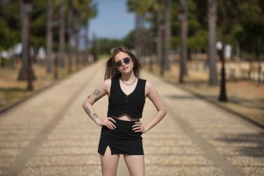 Genç bir kadın, güzel, sarışın, siyah bluzlu, etekli ve güneş gözlüklü, elleri belinde, güçlü ve bağımsız, bir caddenin ortasında. Kadınlık kavramı, güçlenme..