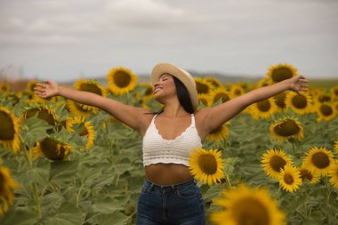 Genç, güzel, Latin esmer kadın beyaz örgü örer, kot pantolon giyer ve şapka giyer, kolları açık, minnettar ve mutlu bir şekilde ayçiçeği tarlasının ortasında. Kavram: doğa, tarım.
