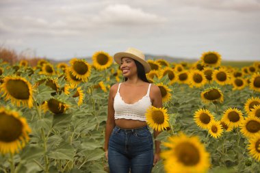 Genç, çekici, latin esmer kadın beyaz örgü örer, kot pantolon giyer ve şapka giyer. Ayçiçeği tarlasının ortasında gülümser ve mutlu olur. Kavram: doğa, tarım, kırsal.
