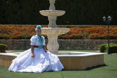 Genç, güzel, sarışın bir kadın tipik Endülüs flamenko kostümlü açık hava parkındaki güzel bir çeşmede açık mavi şallı ve çiçekli bir hac yolculuğu için bekliyor..