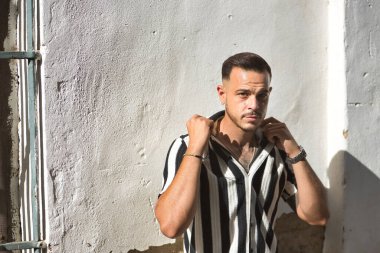 Genç, esmer, İspanyol, modern adam, siyah beyaz çizgili tığ işi gömlek giyiyor, flörtleşerek gömlek yakalarını kaldırıyor, beyaz bir duvarda güneş ışığı alıyor..