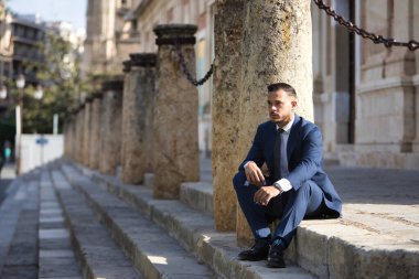 Genç, esmer, İspanyol, modern yönetici, takım elbiseli ve kravatlı, ciddi, ekonomik krizden endişeli ve sıkıntılı, bir şehir caddesinde bazı basamaklarda oturuyor. İş konsepti, şirket, lider.