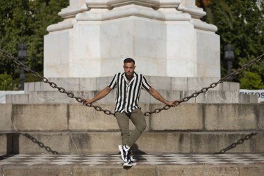 Genç, esmer, İspanyol, modern adam, siyah beyaz çizgili tığ işi gömlek ve kot pantolon giyiyor, ciddi ve cüretkar bakışlı, kocaman bir zincirin üzerinde oturuyor. Moda, moda, gündelik konsept.