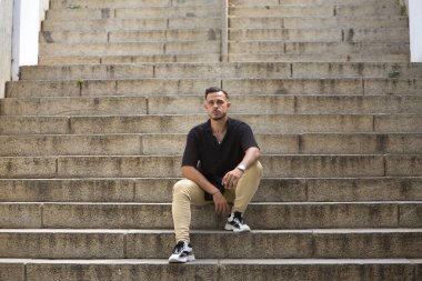 Genç, esmer, İspanyol, siyah tığ işi gömlek ve kot pantolon giyen, yalnız bir merdivenin ortasında sigara içen modern bir adam. Moda, moda, gündelik konsept.