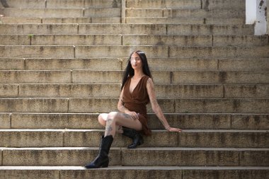 Genç bir kadın, esmer, uzun boylu, zayıf ve çok dövmeli, kahverengi elbiseli ve çizmeli, tatlı, masum ve yalnız, buharlaşmış, merdivenlerde oturuyor. Gençlik kavramı, güzellik, asilik, milenyum.