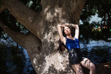 Genç bir kadın, koyu saçlı, uzun boylu, zayıf ve çok dövmeli, mavi bir bluz ve deri şort giymiş, güneşi emen büyük bir ağacın gövdesine yaslanmış. Gençlik kavramı, güzellik, milenyum.