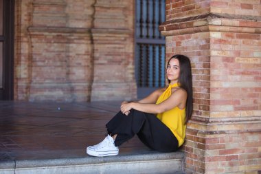 Genç bir kadın, esmer, Latin, sarı gömlek, siyah kot pantolon ve beyaz spor ayakkabı giyiyor, sakin ve rahat, tuğla duvara yaslanmış. Kavram: gençlik, moda, moda, modern.