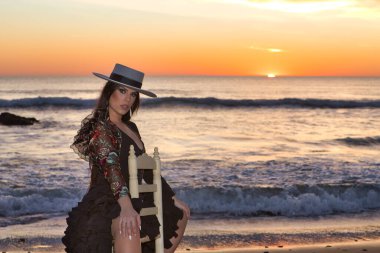 Kadın, genç, güzel, esmer, flamenko dansçısı, kordova şapkalı ve çiçekli siyah flamenko elbiseli, plajda arka planda deniz olan bir sandalyede oturuyor..