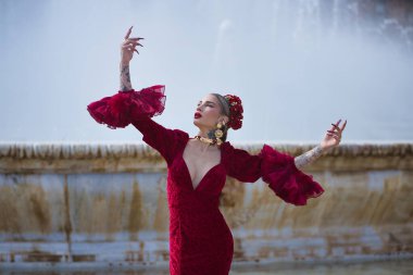 Genç, güzel, sarışın, kırmızı flamenko elbiseli, saçında çiçekler, altın mücevherler, Seville 'de İspanya meydanında fıskiyeli fıskiyelerle flamenko pozları veriyor..