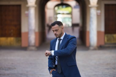 Genç, esmer, İspanyol, modern yönetici, takım elbiseli ve kravatlı, bir sonraki buluşmaya yetişmek için saate bakıyor, eli cebinde, ıssız bir şehir sokağında. Konsept iş, şirket.