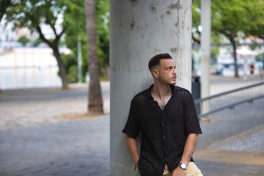 Genç, esmer, İspanyol, siyah tığ işi gömlekli, ciddi ve endişeli bir şekilde poz veren, el ele verip bir sütuna yaslanan modern bir adam. Moda, moda, gündelik konsept.