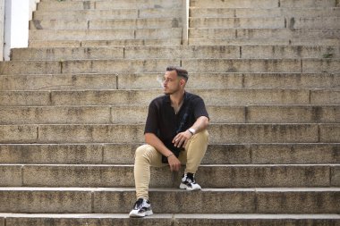 Genç, esmer, İspanyol, siyah tığ işi gömlek ve kot pantolon giyen, yalnız bir merdivenin ortasında sigara içen modern bir adam. Moda, moda, gündelik konsept.