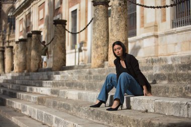 Genç bir kadın, çekici, esmer, mavi gözlü, İspanyol, siyah ceket giyen, kot giyen, topuklu giyen, şehir merkezinde merdivenlerde otururken poz veren. Konsept: model, güzellik, moda, moda.