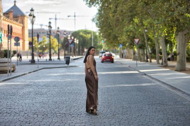 Genç bir kadın, güzel, esmer, mavi gözlü, İspanyol, zarif kahverengi bir şifon elbise ve altın topuklu ayakkabı giyiyor, kentteki kaldırımlı bir sokakta yürüyor. Konsept: model, güzellik, moda.