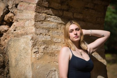 Genç, güzel, sarışın, siyah gömlekli, masum ve hassas bir kadının portresi. Kameraya bakıyor, tuğla kemere yaslanıyor, açık hava parkında güneş ışınları alıyor. Güzellik kavramı, gençlik.