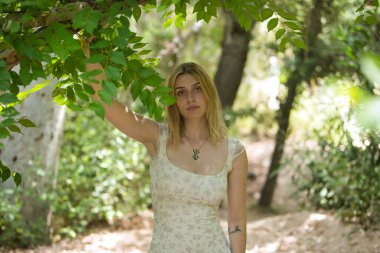 Genç, güzel, sarışın bir kadın, çiçekli bir elbise giyiyor, tatlı ve şefkatli bir bakışla, bir ağacın dalına tutunuyor, doğa ve ekolojiyle bağlantılı, açık hava parkında. Güzellik kavramı, gençlik.