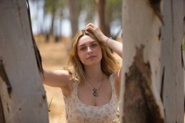 Genç bir kadın, güzel, sarışın, çiçekli bir elbise giymiş, gülümseyen ve mutlu, açık hava parkındaki bir ağacın gövdesine yaslanmış kameraya bakıyor. Güzellik kavramı, gençlik, milenyum.