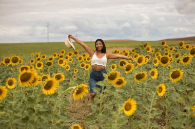 Genç, çekici, latin esmer kadın beyaz örgü örer, kot pantolon giyer ve elinde bir şapka, kolları ayçiçeği tarlasında mutlu bir şekilde gülümserdi. Kavram: doğa, tarım.