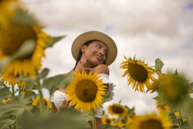 Beyaz bir silindir ve şapka giyen güzel Latin esmer bir kadının portresi, ayçiçeği tarlasında bitkilerin arasında kendini kucaklıyor ve seviyor. Kavram: doğa, tarım, kırsal.
