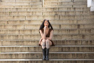 Genç bir kadın, koyu saçlı, uzun boylu, zayıf ve çok dövmeli, kahverengi bir elbise ve çizme giyiyor, eli çenesinde, kameraya bakıyor, tatlı, masum ve yalnız, merdivenlerde oturuyor..