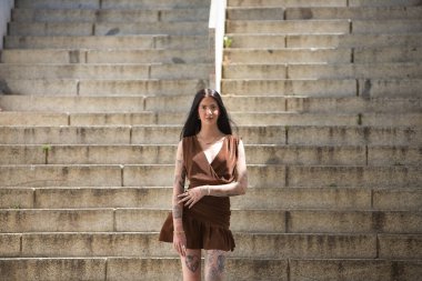 Genç bir kadın, esmer, uzun boylu, zayıf ve çok dövmeli, kahverengi bir elbise giyiyor, kameraya bakarken poz veriyor, tatlı, masum ve yalnız, merdivenlerde duruyor. Gençlik kavramı, bin yıllık.