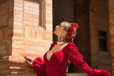 Genç, güzel, sarışın bir kadının portresi, kırmızı flamenko elbiseli, saçında çiçekler ve altın mücevherlerle, İspanya meydanında flamenko pozları veriyor. Dans konsepti, tipik İspanyol..