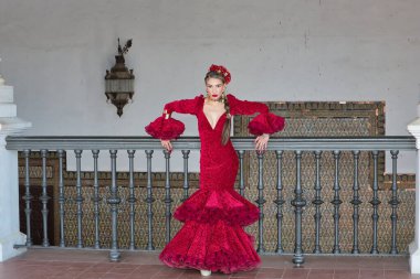 Genç, güzel, sarışın, kırmızı flamenko elbiseli, saçında çiçekler ve altın mücevherlerle, Seville 'de İspanya meydanında metal bir korkuluğa yaslanmış flamenko pozları veriyor. Dans konsepti, sanat.