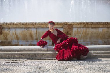 Genç, güzel, sarışın, kırmızı flamenko elbiseli, saçında çiçekler ve altın mücevherlerle, İspanya 'nın Seville meydanındaki güzel bir çeşmede flamenko pozları veriyor. Dans konsepti, sanat.