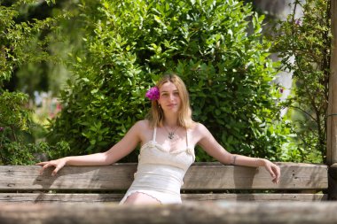 Genç, güzel, sarışın, beyaz elbiseli, kulağında menekşe çiçekleri olan, gülümseyen ve mutlu, açık hava parkındaki ahşap bankta oturan bir kadın. Güzelliği kavra, gençliği, milenyumu.
