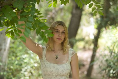 Genç, güzel, sarışın bir kadın, çiçekli bir elbise giyiyor, tatlı ve şefkatli bir bakışla, bir ağacın dalına tutunuyor, doğa ve ekolojiyle bağlantılı, açık hava parkında. Güzellik kavramı, gençlik.