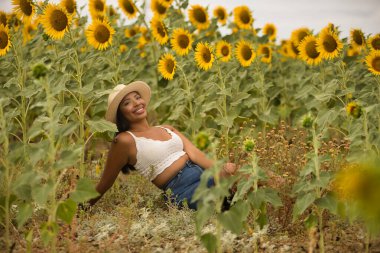 Genç, çekici, latin esmer kadın beyaz örgü örer, kot pantolon giyer ve şapka giyer, ayçiçeği tarlasında mutlu bir şekilde gülümser. Kavram: doğa, tarım, kırsal.