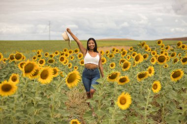 Genç, çekici, latin esmer kadın beyaz örgü örer, kot pantolon giyer ve elinde bir şapka, kolları ayçiçeği tarlasında mutlu bir şekilde gülümserdi. Kavram: doğa, tarım.