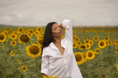 Genç, güzel, Latin esmer kadın beyaz gömlek giyiyor, gözleri kapalı, doğaya bağlı, ayçiçeği tarlasının ortasında. Kavram: doğa, tarım, kırsal.