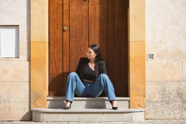 Genç bir kadın, çekici, esmer, mavi gözlü, İspanyol, siyah ceketli ve bluzlu, kot pantolonlu, güzel bir kapının eşiğinde otururken poz veren. Konsept: model, güzellik, moda, moda.