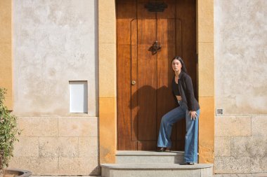 Genç bir kadın, çekici, esmer, mavi gözlü, İspanyol, siyah ceket ve bluz giyiyor, kot pantolon giyiyor, kameraya bakıyor, kapının çerçevesine yaslanıyor. Konsept: model, güzellik, moda.