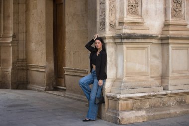 Genç bir kadın, çekici, esmer, mavi gözlü, İspanyol, siyah ceket ve bluz giyen, kot giyen, topuklu giyen, şehir merkezinde duvara yaslanmış poz veren. Konsept: model, güzellik, moda, moda.