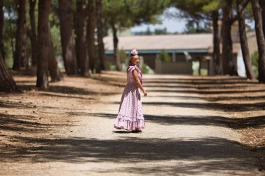 Genç bir kadın, güzel, sarışın, tipik Endülüs flamenko kostümlü, küçük şallı ve pembe çiçekli, kırsalda tek başına geriye bakan..