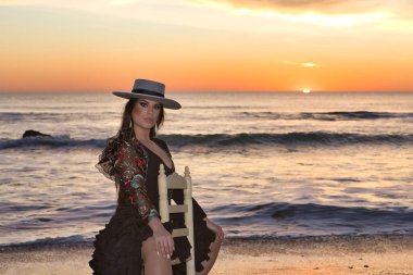 Kadın, genç, güzel, esmer, flamenko dansçısı, kordova şapkalı ve çiçekli siyah flamenko elbiseli, plajda arka planda deniz olan bir sandalyede oturuyor..