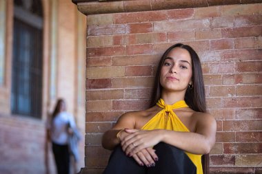 Genç, koyu renk saçlı Latin bir kadının portresi sarı gömlek ve siyah kot pantolon giymiş, tatlı ve hassas, kameraya bakıyor ve tuğla duvara yaslanıyor. Kavram: Gençlik, moda.