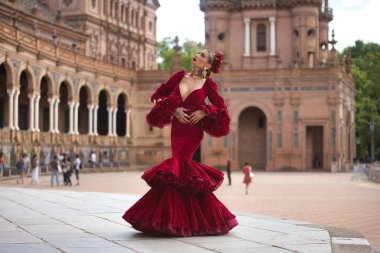 Genç, güzel, sarışın, kırmızı flamenko elbiseli, saçında çiçekler ve altın mücevherlerle İspanya meydanında flamenko pozları veriyor. Dans konsepti, sanat, tipik İspanyol..
