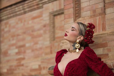 Genç, güzel, sarışın bir kadının portresi, kırmızı flamenko elbiseli, saçlarında çiçekler ve altın mücevherler, hislerle kapalı gözler, Seville 'de İspanya meydanında bir tuğla duvara yaslanmış..