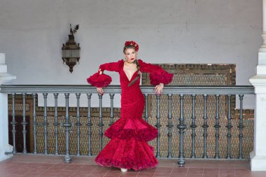 Genç, güzel, sarışın, kırmızı flamenko elbiseli, saçında çiçekler ve altın mücevherlerle, Seville 'de İspanya meydanında metal bir korkuluğa yaslanmış flamenko pozları veriyor. Dans konsepti, sanat.