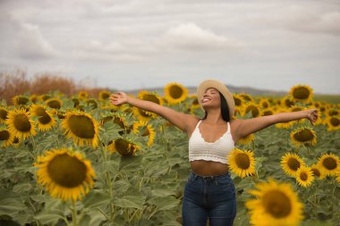 Genç, güzel, Latin esmer kadın beyaz örgü örer, kot pantolon giyer ve şapka giyer, kolları açık, minnettar ve mutlu bir şekilde ayçiçeği tarlasının ortasında. Kavram: doğa, tarım.