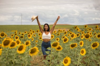 Genç, çekici, latin esmer kadın beyaz örgü örer, kot pantolon giyer ve elinde bir şapka, kolları ayçiçeği tarlasında mutlu bir şekilde gülümserdi. Kavram: doğa, tarım.