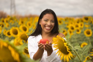 Genç, çekici, Latin esmer kadın beyaz gömlek giyiyor, mutlu bir şekilde gülümsüyor ve ayçiçeği tarlasının ortasında bir elma tutuyor. Kavram: doğa, tarım, kırsal.