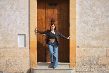 Genç bir kadın, güzel, esmer, mavi gözlü, İspanyol, siyah ceket ve bluz giyiyor, kot pantolon giyiyor, elleri güzel bir kapının çerçevesinde duruyor. Konsept: model, güzellik, moda, moda.