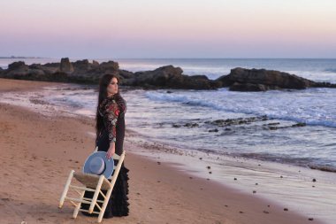 Kadın, genç, güzel, esmer, flamenko dansçısı, çiçekli siyah bir flamenko elbisesi var, elinde sandalye ve elinde bir Cordova şapkasıyla sahilde yürüyor, arkasında deniz var..