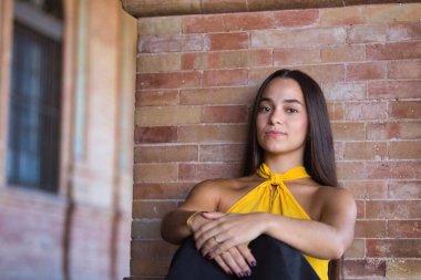 Genç, koyu renk saçlı Latin bir kadının portresi sarı gömlek ve siyah kot pantolon giymiş, tatlı ve hassas, kameraya bakıyor ve tuğla duvara yaslanıyor. Kavram: Gençlik, moda.