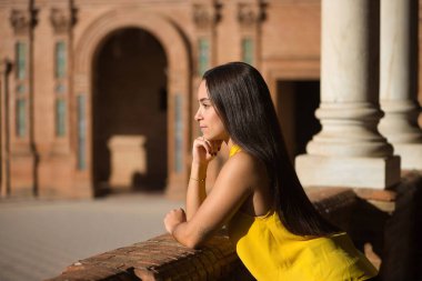 Genç bir kadın, esmer, Latin, sarı tişörtlü, tuğla parmaklıklara yaslanmış, Sevilla 'daki İspanya meydanının ihtişamını seyrediyor. Kavram: gençlik, turizm, seyahat, büyüleyici yerler.