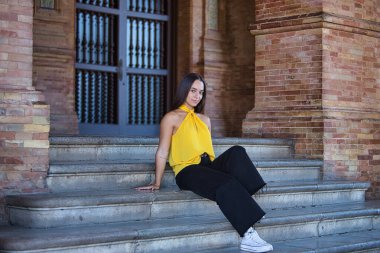 Genç bir kadın, esmer, Latin, sarı gömlek, siyah kot pantolon ve beyaz spor ayakkabı giyiyor, gülümsüyor ve mutlu, kameraya bakıyor, merdivenlerde oturuyor. Kavram: gençlik, moda, moda, modern.